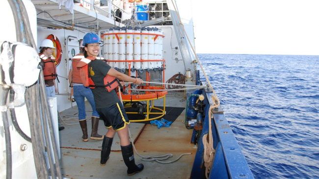 A person posing while working with ropes on a ship. She is wearing a hard hat, a life jacket, and waterproof boots.