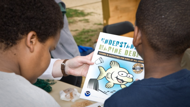 Two children are seen from behind looking at the cover of an activity book featuring a cartoon fish surrounded by marine debris. The cover reads "Understanding Marine Debris: Games and Activities for Kids of All Ages." 