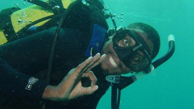 A scuba diver giving the diving "OK" symbol with their fingers. The diver does not have any breathing apparatus in their mouth and is smiling for the photo.