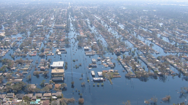 An aerial photograph of a flooded residential area. All roads and ground are covered by water. Houses, buildings, and trees are visible above the flooded areas.