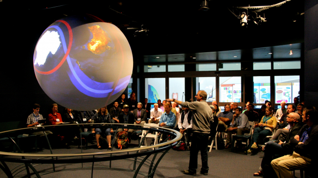A presenter speaking to a group of people, gestoring towards a 6 foot sphere explaining earth science data.