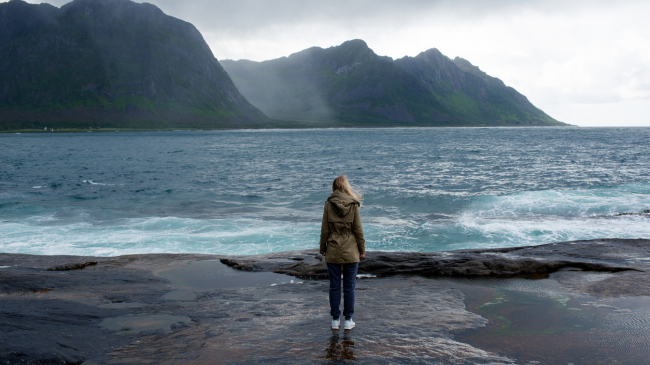 A woman standing on a flat rock ledge of a beach looks out at the blue ocean off the coast of Norway. Tall mountains lay in the distance. Undated 2019 image.