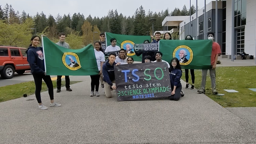 A group of students stand outside their school while holding a sign that reads "TSSO Tesla STEM Science Olympiad Nats 2022."