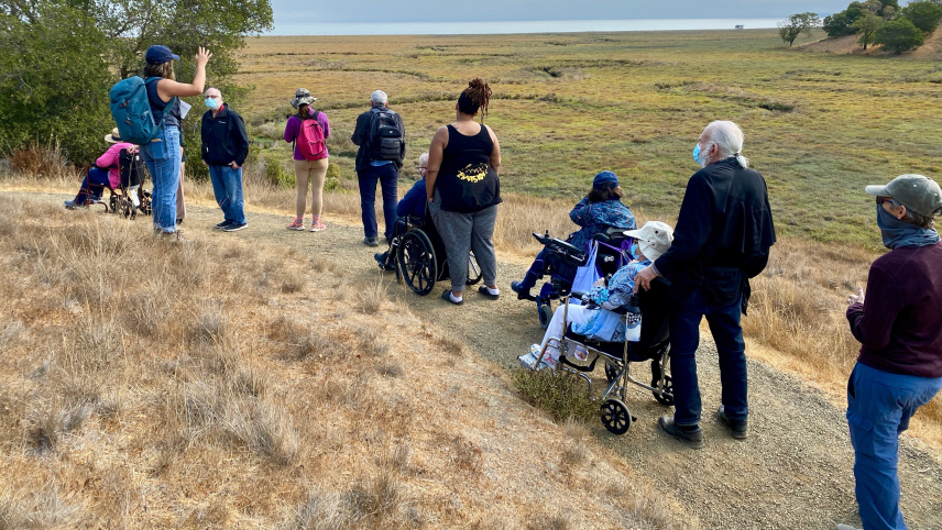 Bella Mayorga speaks and gestures to a group of 10 people, four of whom are in wheel chairs. They are stopped on a path on a grassy hill overlooking salt marshes, oak trees, and a view of a bay.