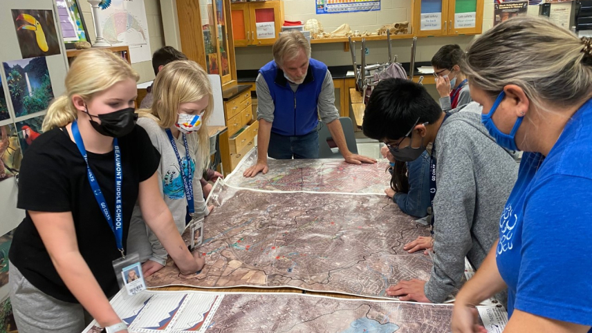 Patrick Geoff, his students, and another adult stand around a table to look at several large city maps. 
