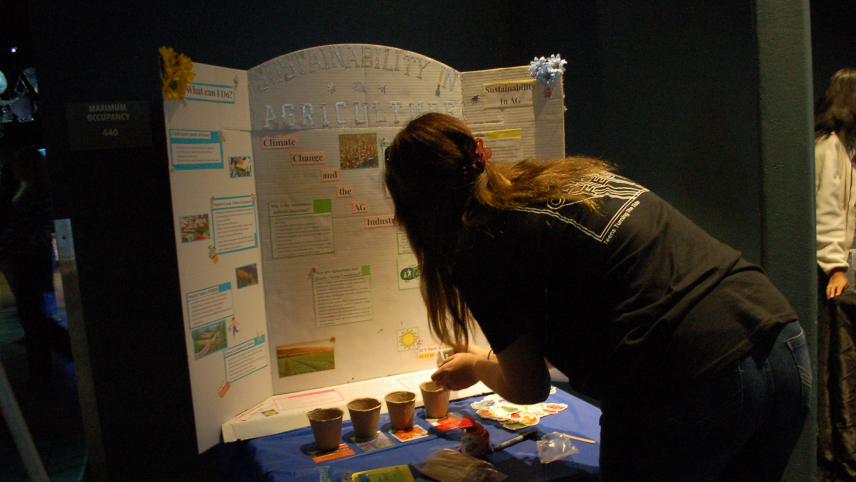 A person is pouring something into one of four small pots that sit on top of a table. A handmade poster titled “Sustainability in Agriculture” is also displayed.