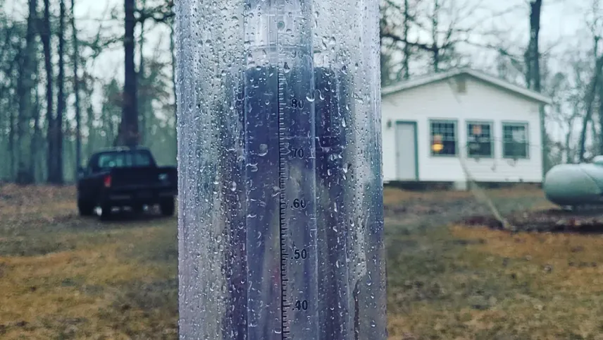 A wide clear plastic tube, sitting in front of a white house and black pickup truck with colorful leaves strewn across the ground, is covered in what appears to be rain and has a smaller tube within it measuring the water collected.