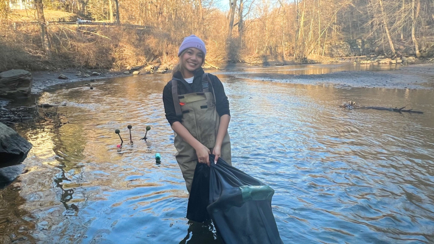 Eva Lagdamen is staning in a river with relatively still water holding a large black nylon bag that is used to capture glass eels for counting. She is surrounded by a few rocks, as well as bare brush in the background. She is wearing a purple beanie, a black sweatshirt, and brown waders.