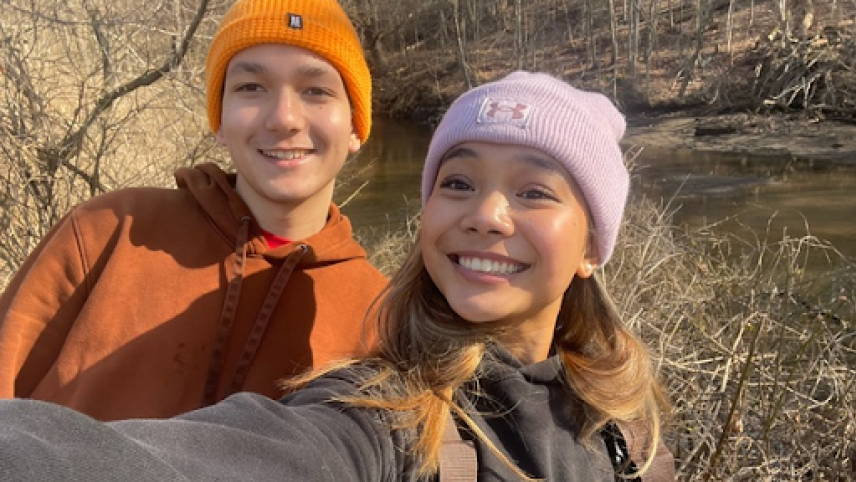 Eva and Benjamin are posing together for a selfie on the side of the road, overlooking the river. It is early spring, and the brush around them has not yet started to bloom. Benjamin is wearing a bright orange beanie as well as a muted orange sweatshirt. Eva is wearing a purple beanie, a black hoodie, an brown waders.