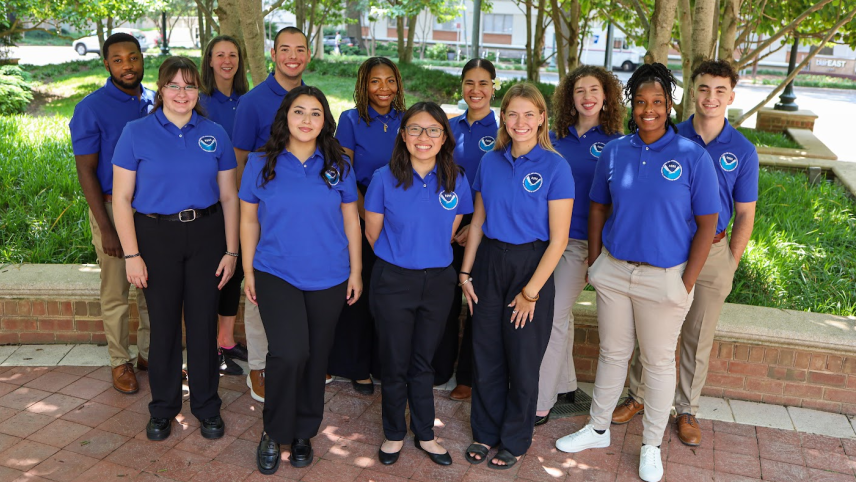 Twelve people pose and smile for the camera outside. They are wearing blue polo shirts that have a NOAA logo in the upper right. 