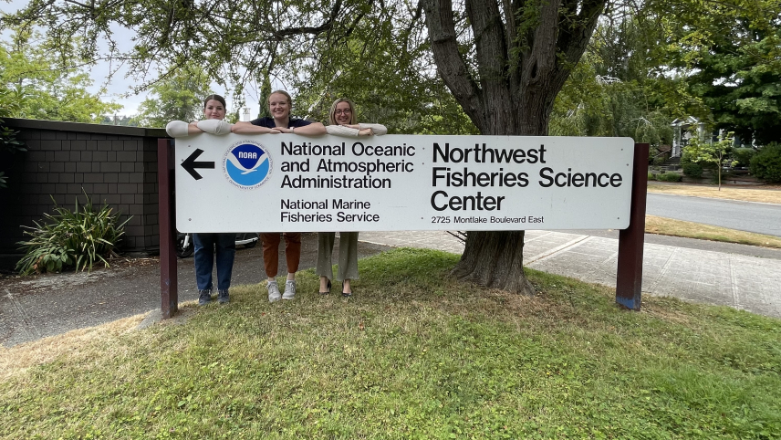 Three women smile, standing behind a monument sign that says, "NOAA Fisheries Northwest Fisheries Science Center."