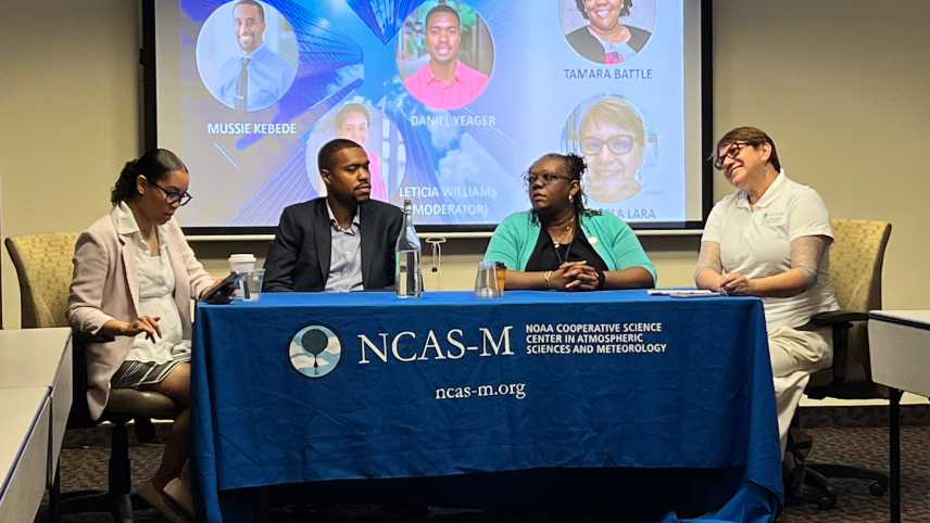 A panel of four people who are sitting at a table with an NCAS-M table cloth. Behind them is a projector screen, with a slide titled "Interdisciplinary science alumni panel."