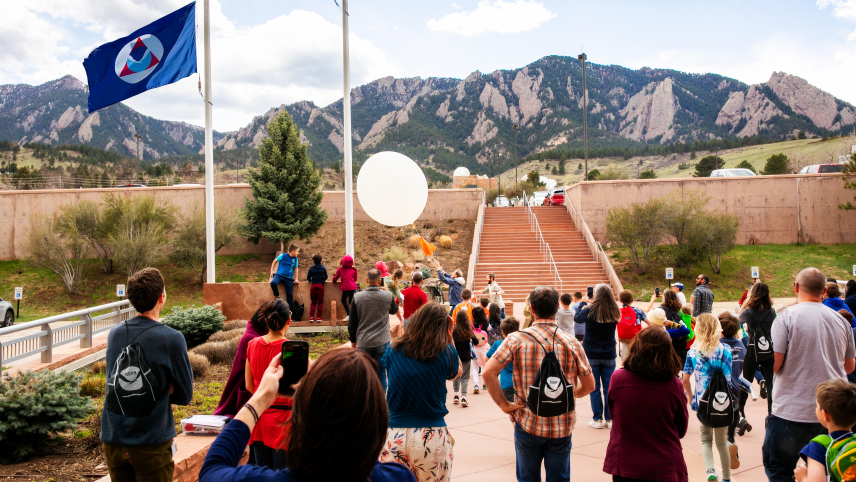Children and adults gather to assist two scientists launching a weather balloon in windy conditions. The U.S., Colorado, and NOAA flags flutter in the breeze, framed by the Flatiron Mountains in the distance.