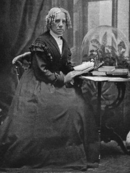 A black and white photo of Maria Mitchell sitting at a small table with an open book.