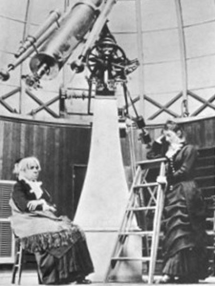 A black and white photo of Maria Mitchell sitting by a large telescope in the Vassar College Observatory with her student Mary Whitney. Circa 1877.