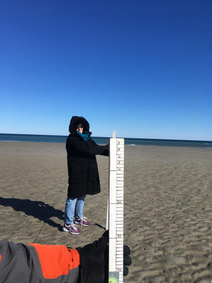  Linda Stathoplos is standing in front of her husband, John, as he snaps a picture showing both her and the measuring stick he uses to collect data on sand contours on the beach. The stick itself is white and has several notches indicating the length, much like a ruler. Linda is wearing a large black jacket with her hood on, blue jeans, and purple tennis shoes. As they stand on the beach, it is a sunny day with the sea in the background.