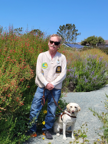 Docent Ron Peterson and his guide dog Gidget stand near a large bush  covered in many red, tube-shaped flowers. Behind them, a gravel path winds through more colorful flowers, and the roof of a building with solar panels is visible in the distance.