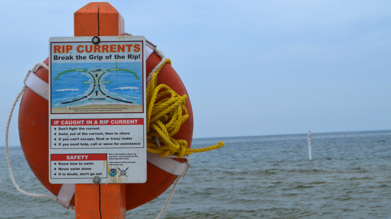 A sign mounted to a bright orange post (which also holds a life ring) is in the foreground of a large body of water. Sign text: Rip currents: Break the Grip of the Rip! If caught in a rip current, don't fight the current. Swim out of the current, then to shore. If you can't escape, float or tread water. If you need help, call or wave for assistance. Safety: Know how to swim. Never swim alone. If in doubt, don't go out.