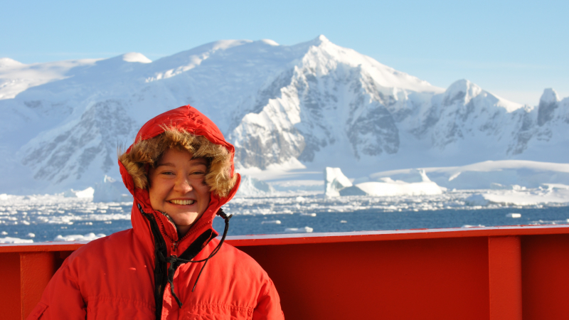Alexa Sterling stands on the deck of a ship wearing a cold weather survival suit. Ice bergs and snow covered mountains are in the background.  