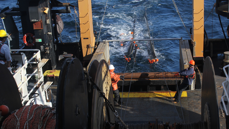 At the end of the trawl, the lead fisherman (far left) operates the winch controls to slowly haul the net on board. Deck crew members keep the net aligned with the large reel in the foreground.