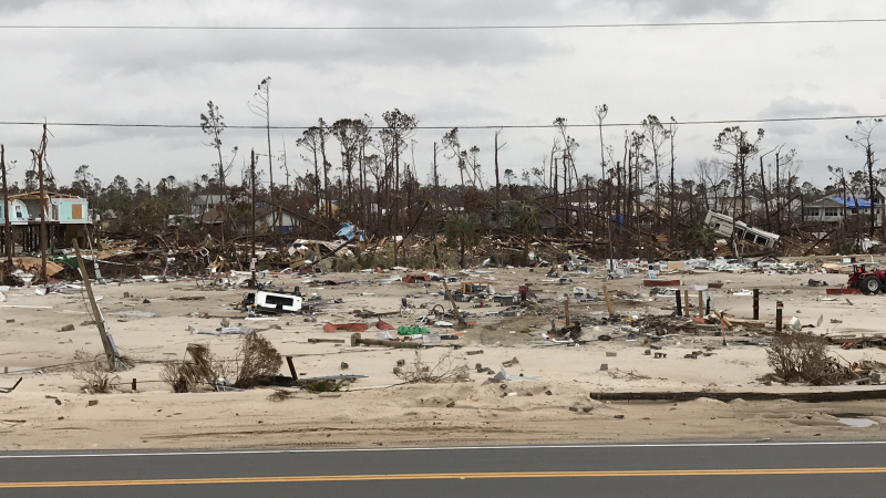 A photo of a mobile home park next to a road. The park has been destroyed due to storm surge. Homes are broken to pieces and there is debris from them everywhere.