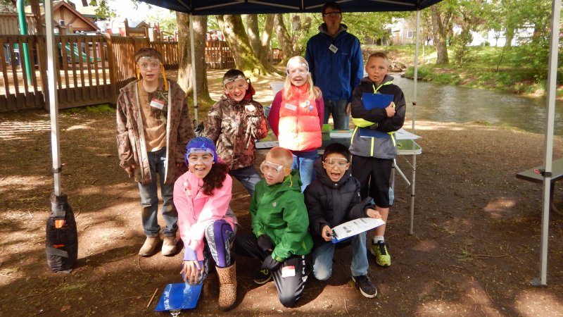 7 students and one staff member look at the camera wearing protective googles underneath a tarp outside. 2 students in the front row are holding species identification keys
