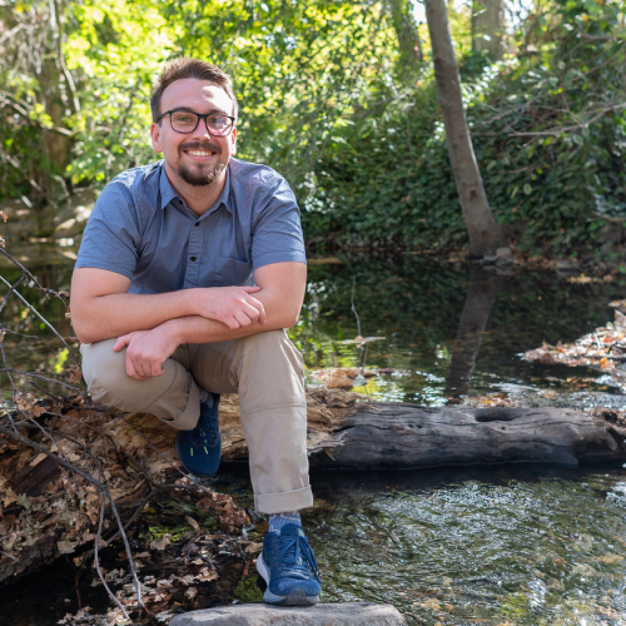 Cole wears business casual clothing and sits on a log in a wooded streamside landscape.