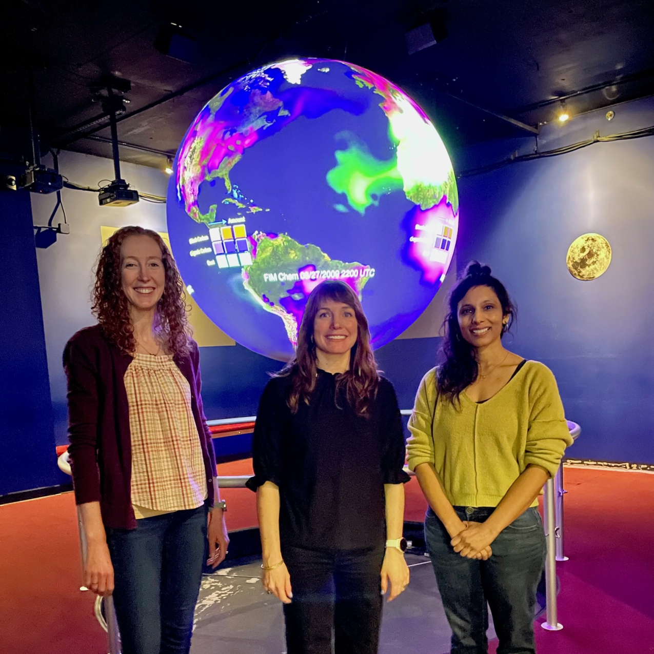 Beth Russell Wehe, Hilary Peddicord, and Shilpi Gupta stand in front of the Science On a Sphere tool, a large globe projector that hangs from the ceiling and projects global science data.