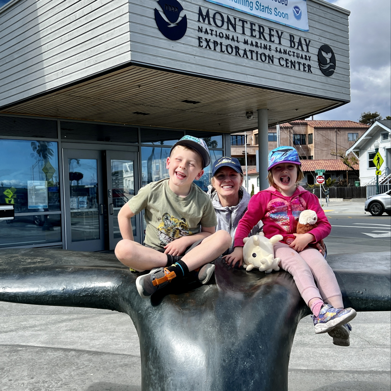 Two children wearing baseball caps sit and pose on a sculpture of a whale’s tale outside in front of a building with a sign that reads “Monterey Bay National Marine Sanctuary Exploration Center.” The child on the right is holding two stuffed animals. An adult wearing a baseball cap stands posing behind them.  