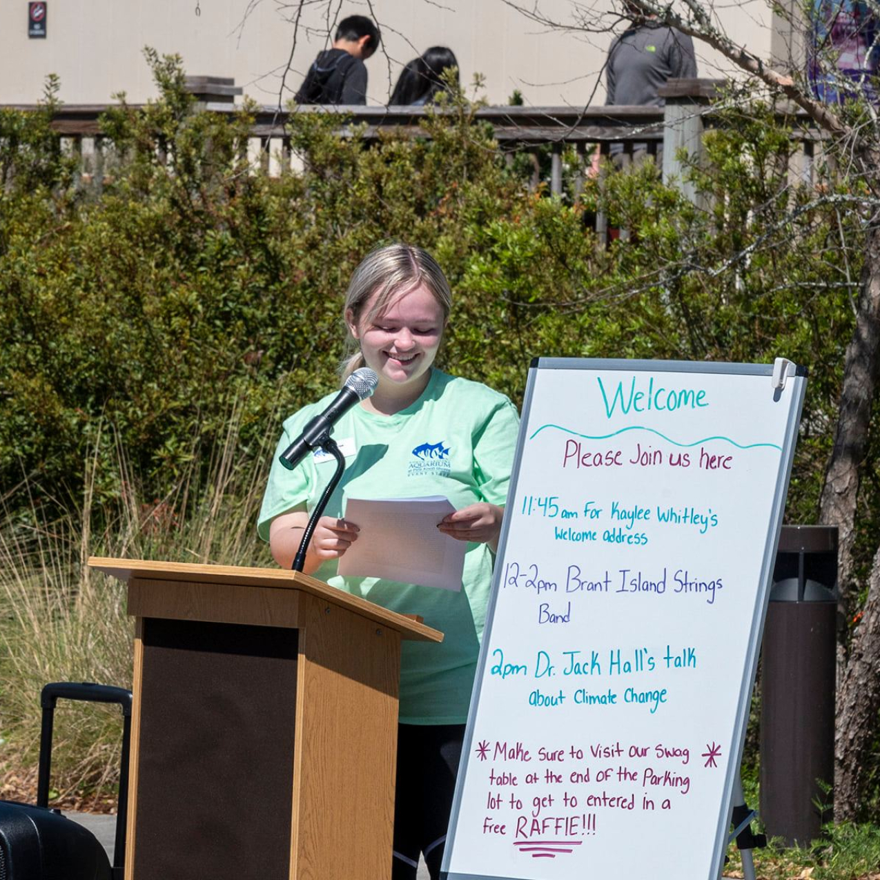 A person stands behind a podium with a microphone outside while holding and looking at papers in her hand. A white board is set up next to her that reads “Welcome please join us here” and lists a schedule of the event. In the background, there are trees and bushes and a few people standing on a wooden deck. 