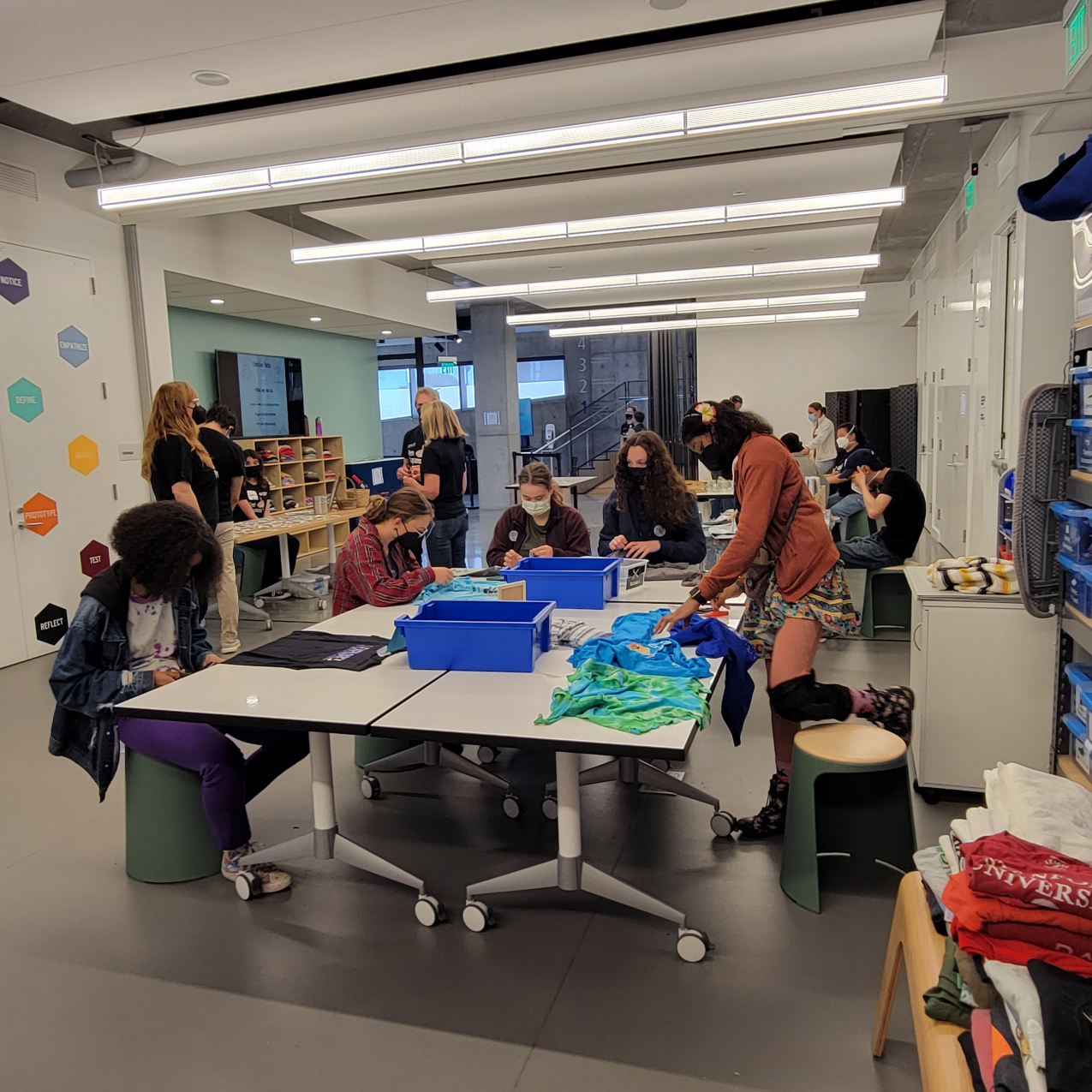 Teens collaborate around a table to work on an activity to upcycle used shirts. There are other groups of people behind them working on different activities at various tables. 