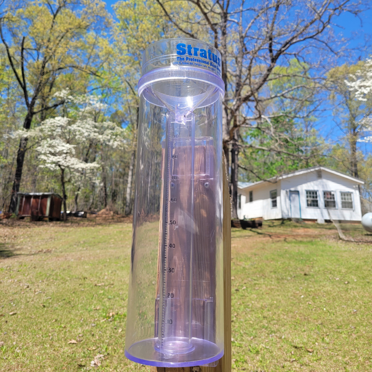 A wide clear plastic tube, sitting in front of a white house with lush grass in the background, basking in sunlight, with no water collected in the smaller measuring tube located within the middle of the large.