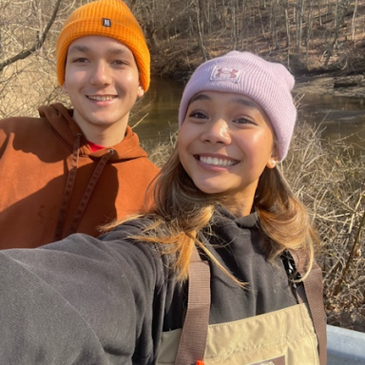 Eva and Benjamin are posing together for a selfie on the side of the road, overlooking the river. It is early spring, and the brush around them has not yet started to bloom. Benjamin is wearing a bright orange beanie as well as a muted orange sweatshirt. Eva is wearing a purple beanie, a black hoodie, an brown waders.