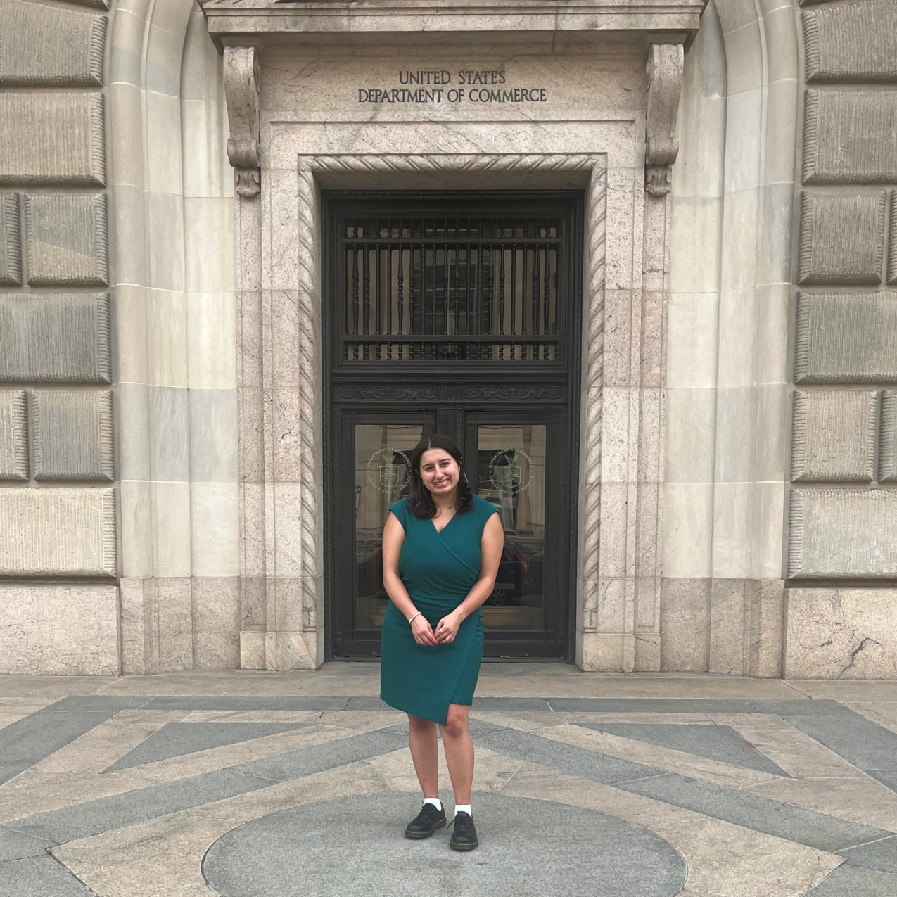 Maria smiles, posing in front of a concrete building with block lettering above the door that reads United States Department of Commerce.