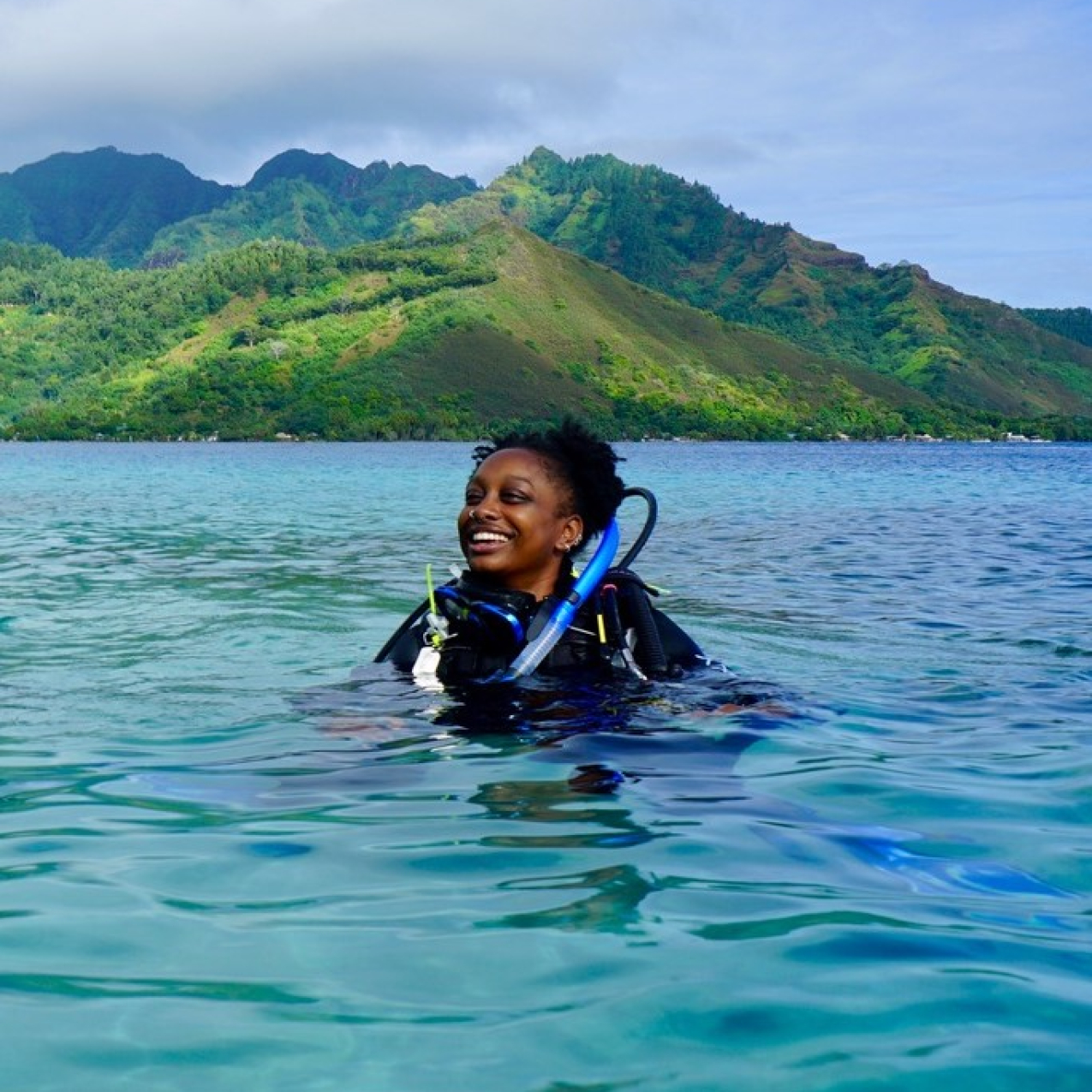 Janelle grins in full SCUBA gear, floating at the surface of crystal clear, blue ocean water. In the background, mountains covered in green vegetation meet the shore.