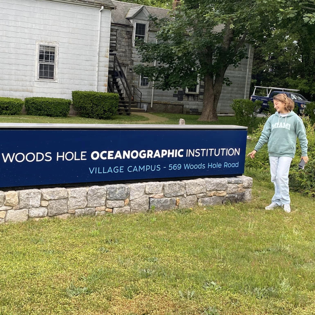Allie stands outside next to a sign that reads "Woods Hole Oceanographic Institution. Village Campus.