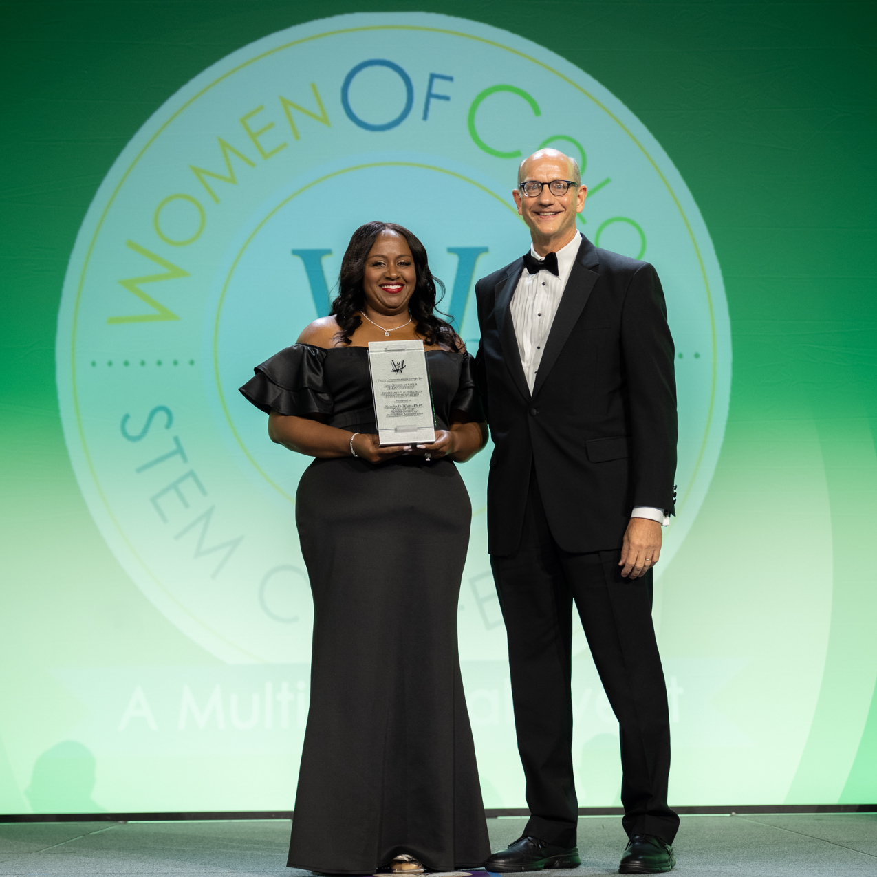 Natasha and the conference host, Eric S. Musser, stand side-by-side in black-tie attire as Natasha accepts a placard as her award. They stand in front of a screen that is projecting the Women of Color in STEM logo.