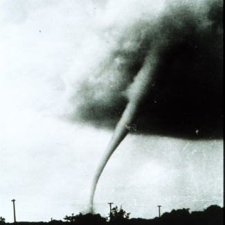 A black and white photo of a tornado moving across a landscape. 