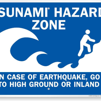 A blue sign with a graphic of a tsunami and a person climbing to higher ground. The text reads: Tsunami Hazard Zone. In case of earthquake, go to high ground or inland