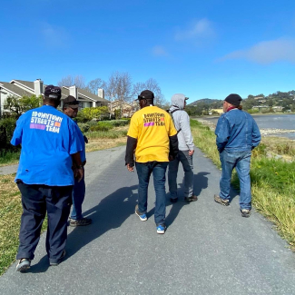 Five people stroll down a paved path. There are houses on one side and a bay on the other. Two people are wearing Downtown Streets Steam t-shirts, and the group appears to be observing the scenery and talking with one another. 