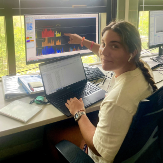 Cassidy is sitting at a work desk, looking at the camera. One of her hands rests on a laptop with data on it, while she uses her other hand to point to a computer monitor displaying multiple waveforms.