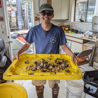 Nick stands in what looks to be a lab for processing field samples. He grins, holding a large tray with oyster shells. His field boots and active transition lenses implied he's just brought them inside.