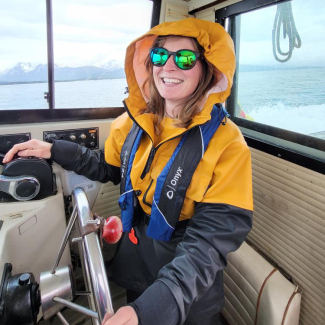 Seraina smiling at the enclosed helm of a boat. She is dressed for fieldwork in cold weather and wears a personal floatation device.