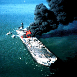 A large barge vessel is on fire in the middle of a blue-green ocean. Big, dark plumes of smoke are coming off of the vessel. There are two small boats that are spraying something on the fire.