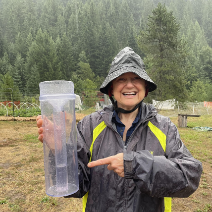 Anne, donned with full rain gear, joyfully holds and points to her filled rain gauge.