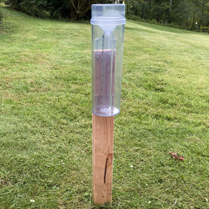 A plastic cylinder with an opening at the top to collect precipitation is mounted upright on a wooden post that stands alone in a grassy field.