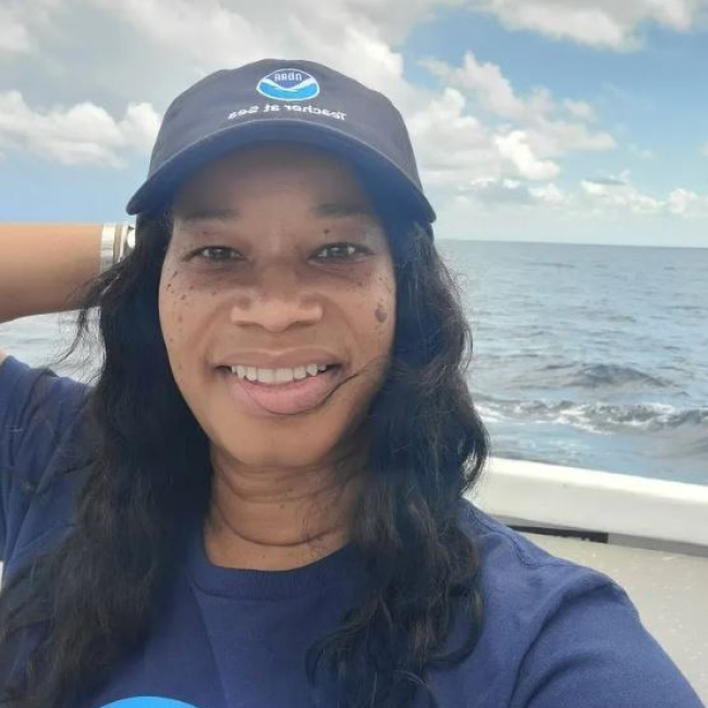Maronda Hastie, wearing a NOAA Teacher at Sea hat, stands on the deck of a ship at sea.