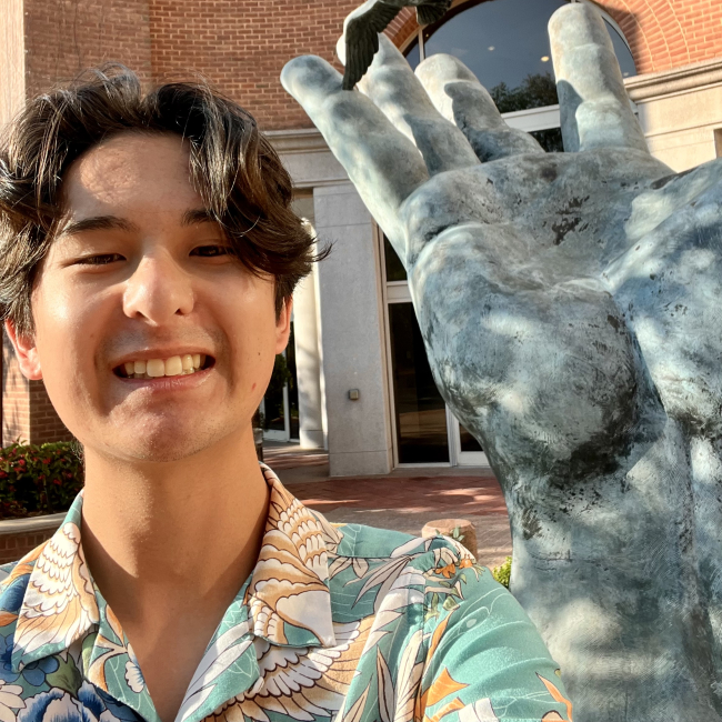 Isaac smiles for a selfie in front of a sculpture of a hand releasing birds, which stands outside of a brick building.