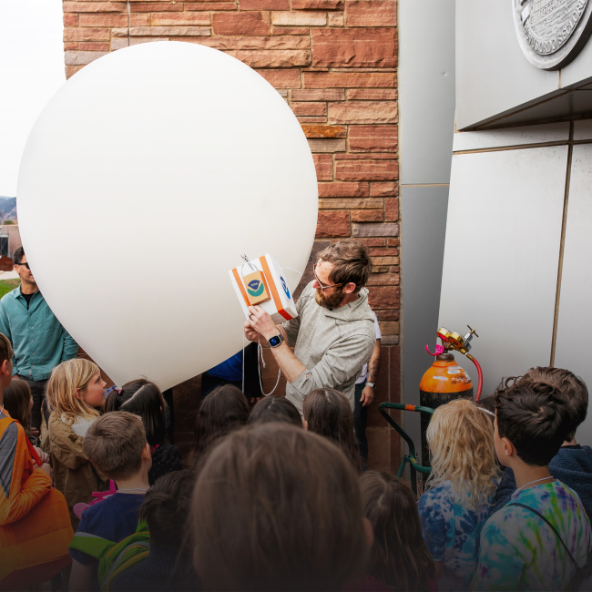 Patrick Cullis, NOAA scientist, surrounded by a sea of enthusiastic children outside the NOAA Boulder building. Boulder's scenic backdrop enhances the enchantment as they eagerly observe the balloon launch, fostering a love for science in young hearts.