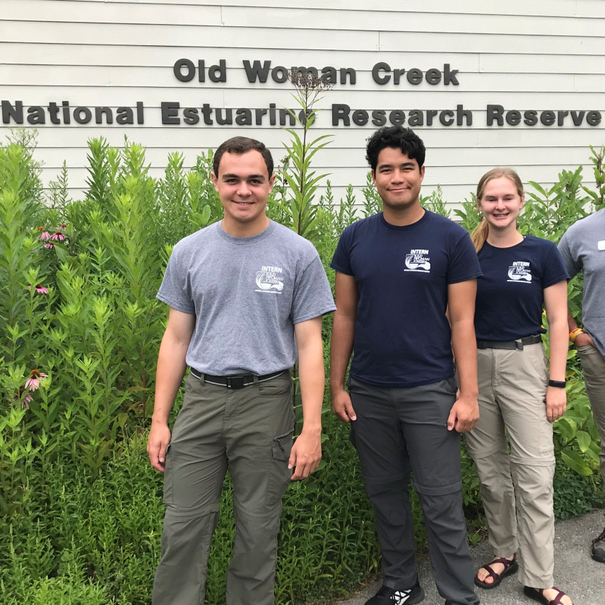 Three young adults in field pants and t-shirts that say "INTERN" above an Old Woman Creek National Estuarine Research Reserve logo pose in front of a pollinator garden and building with block letters that read "Old Woman Creek National Estuarine Research Reserve.